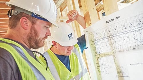 Two men wearing hard hats and other safety gear looking at blue prints