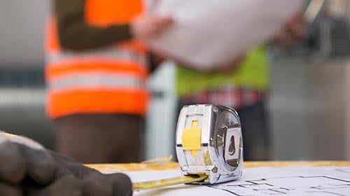 People blurred in the background wearing safety vests with an expanded measuring tape in the foreground