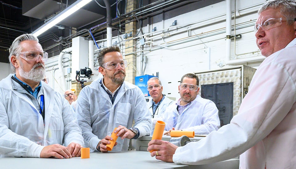 Men in white lab coats standing around a table in a facility holding orange pipe pieces