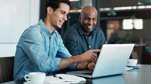 two men in an office setting smiling while looking at something on a laptop screen