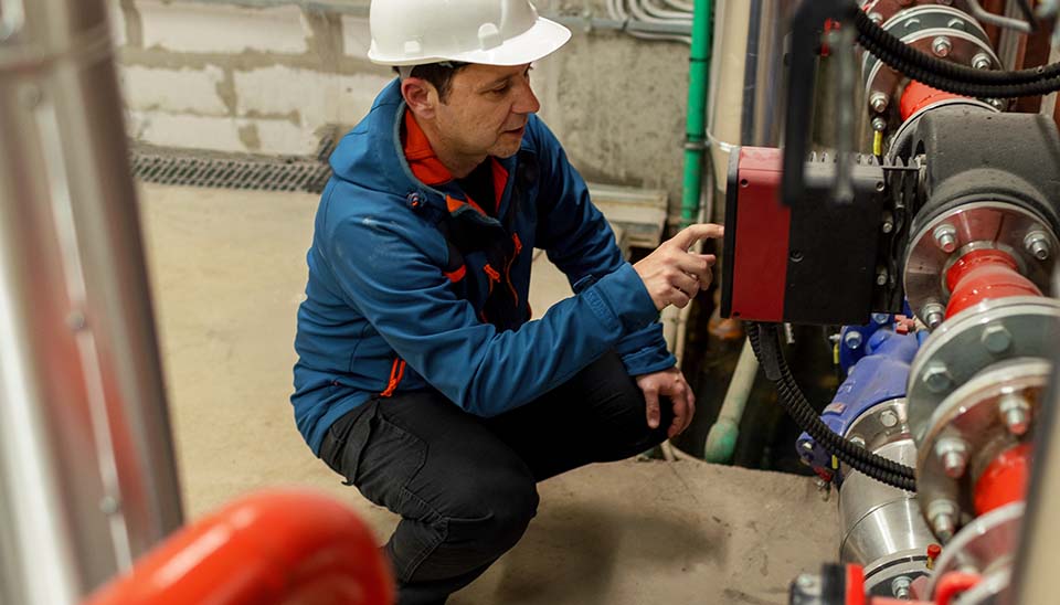 Man wearing white hard hat and blue coat kneeling down while inspecting equipment