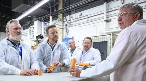 men in lab coats around a table in a warehouse holding plastic piping samples