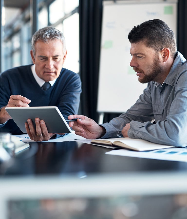 Two men sitting at a table in what looks to be an office setting looking over documents