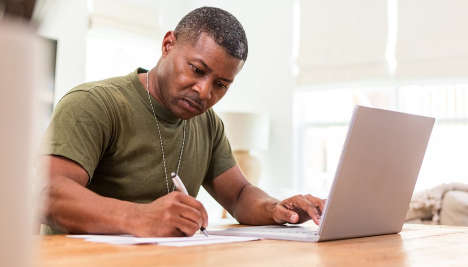 man taking notes on paper while sitting and working at laptop